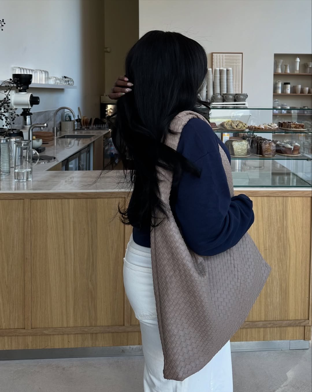 A pessoa tem cabelos compridos, veste blusa azul, calça clara e está de costas em uma cafeteria, segurando uma bolsa feminina bege trançada.