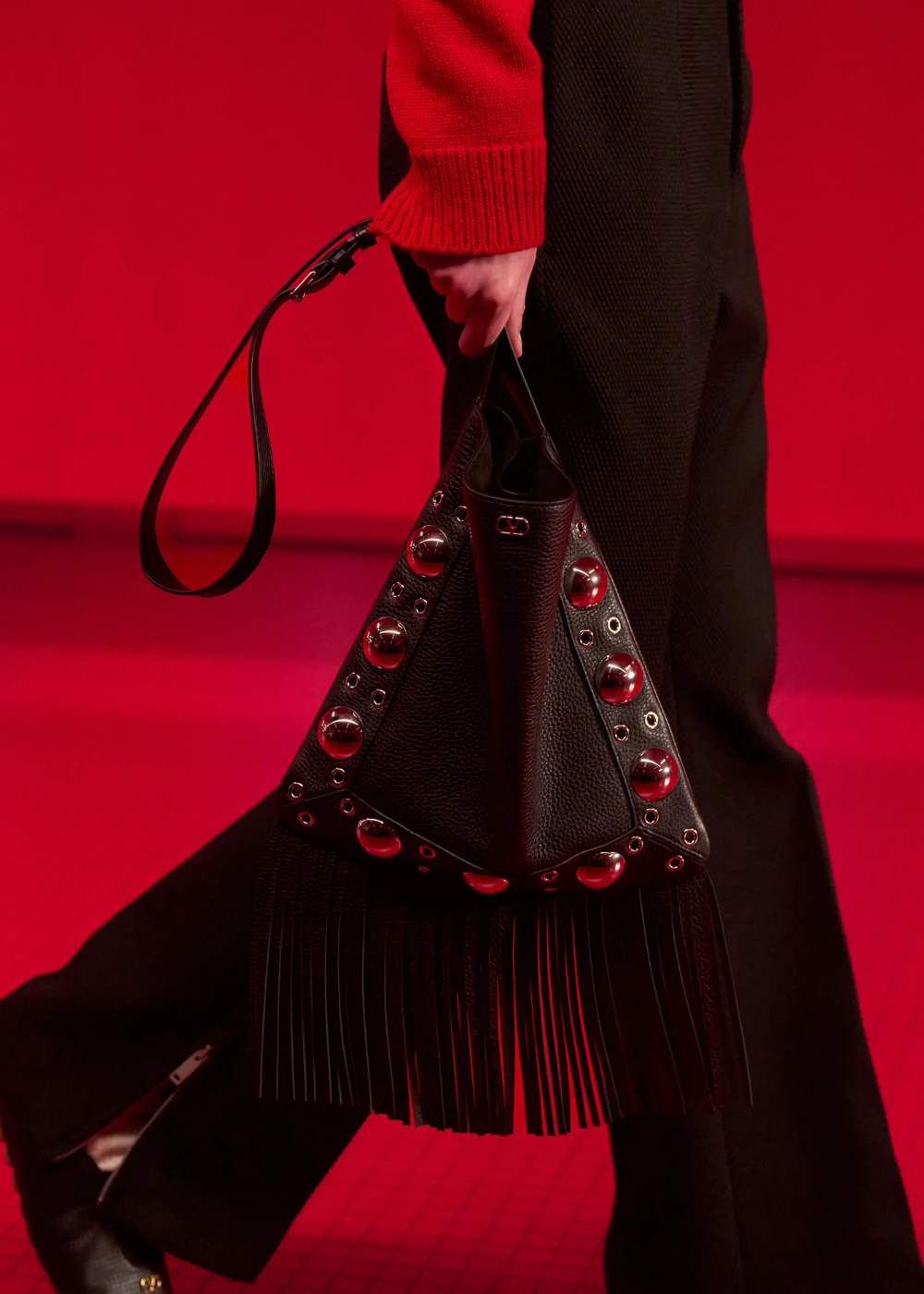 Bolsa triangular preta com franjas e metais, em cenário vermelho, destacando estilo sofisticado.