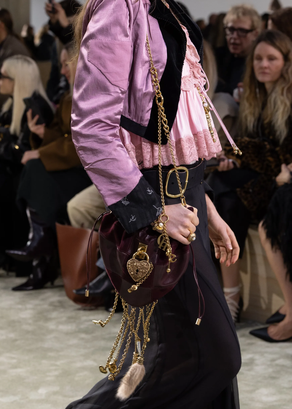 Bolsa feminina estilosa com detalhes dourados em desfile de moda, combinada com roupas em tons de rosa e preto.