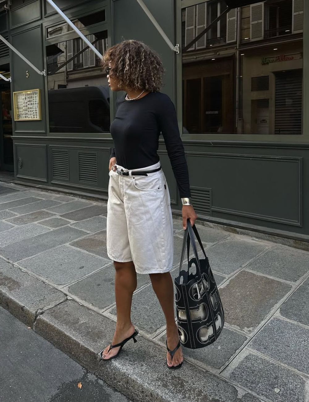 Mulher usando blusa preta e bermuda branca, segurando bolsa estilosa preta e branca na calçada.