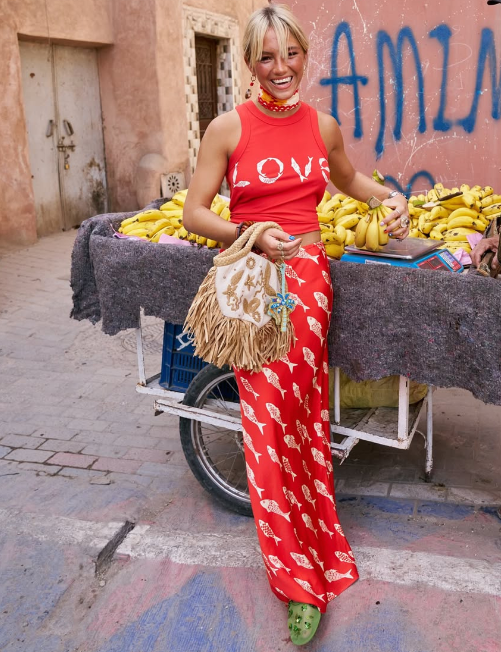 A mulher exibe bolsas femininas estilosas em frente a uma barraca de bananas. Ela sorri, vestida com roupas vibrantes.