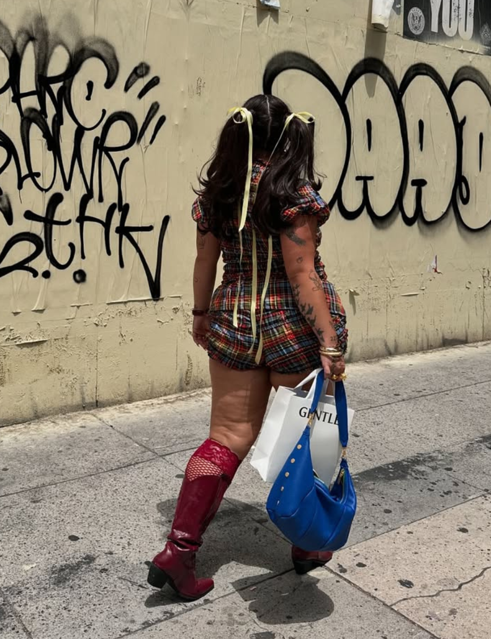 Pessoa andando com botas vermelhas, vestido xadrez e bolsa azul estilosa, em frente a muro com grafite.
