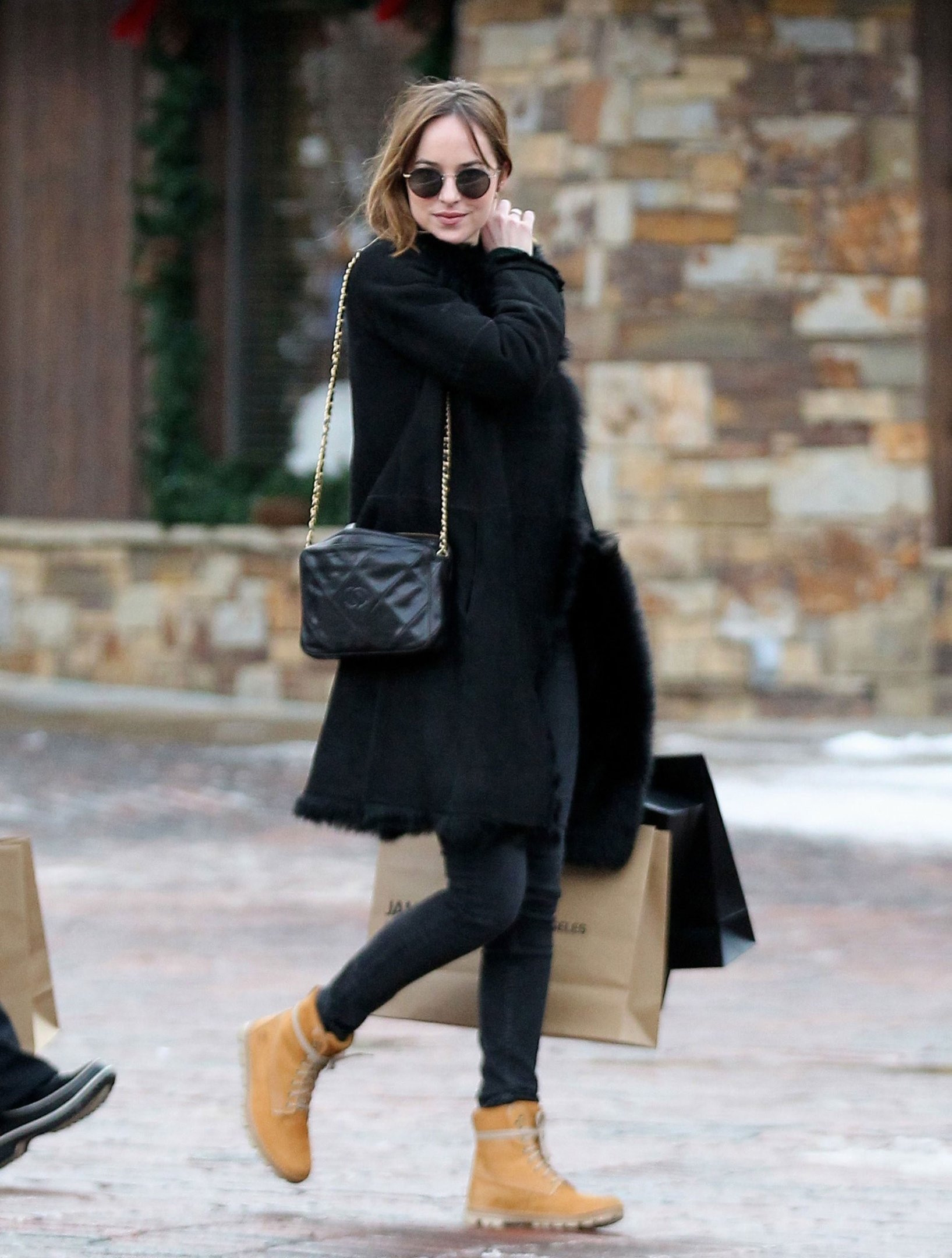 Pessoa usando casaco preto e óculos escuros, com bota Timberland, caminhando na calçada com sacola de compras.