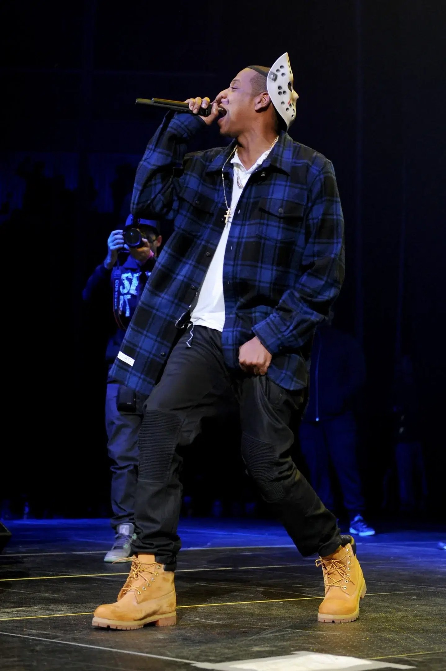 Homem no palco usando camisa xadrez e bota Timberland bege, cantando ao microfone.