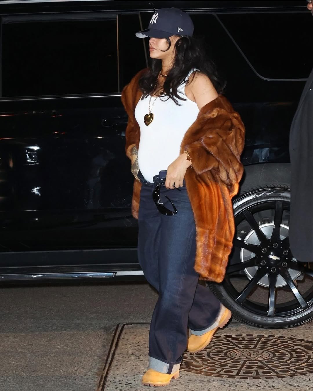 Pessoa vestindo casaco de pele, calça jeans e bota Timberland amarela, perto de um carro preto.
