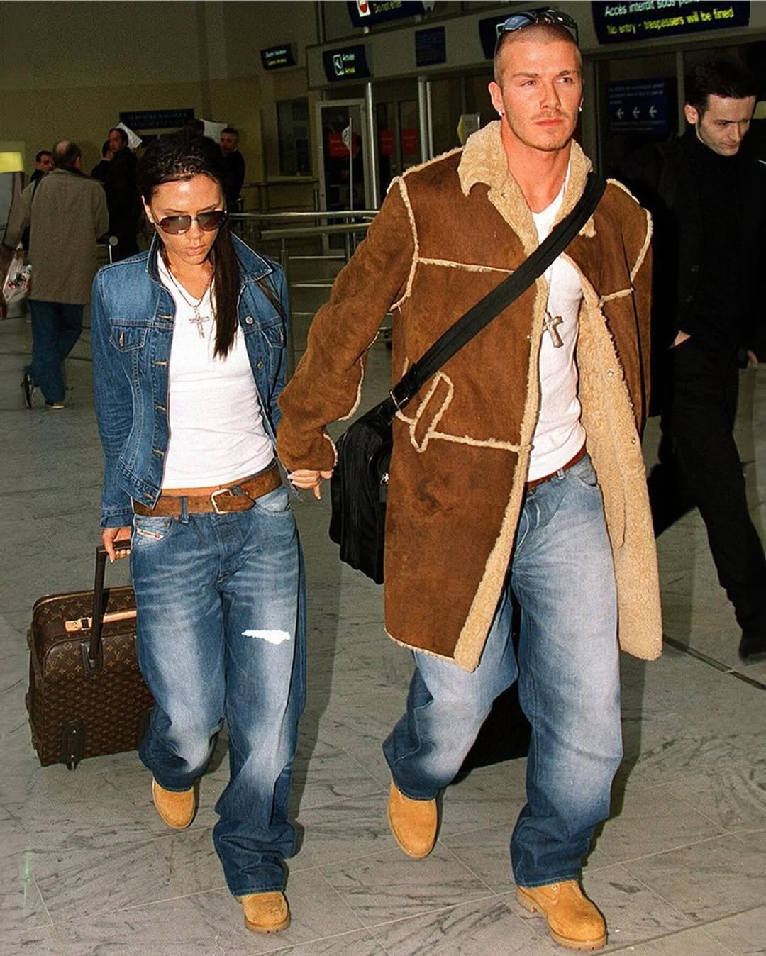 Casal vestindo jaqueta e jeans, caminhando em local público. Ambos usam botas Timberland e seguram malas.