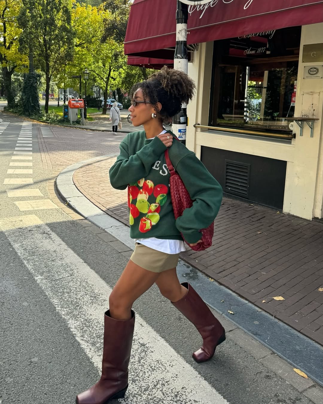 Pessoa caminhando na calçada com suéter verde, bolsa vermelha e botas de bico quadrado em uma cidade arborizada.