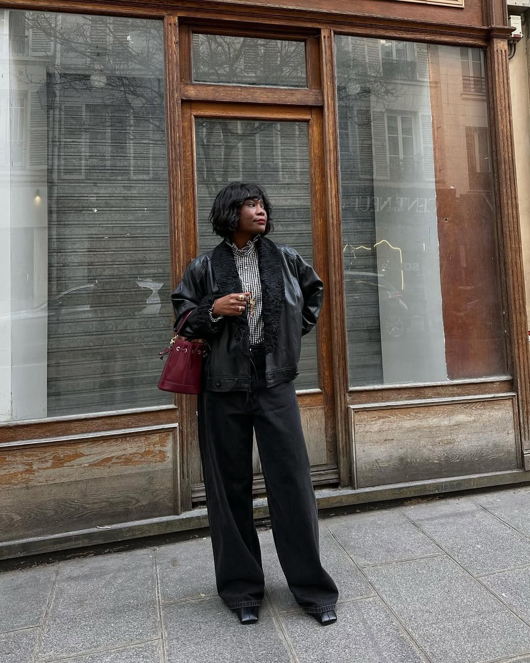 Pessoa em frente a uma vitrine, vestindo calça e jaqueta pretas, camisa listrada e botas de bico quadrado.