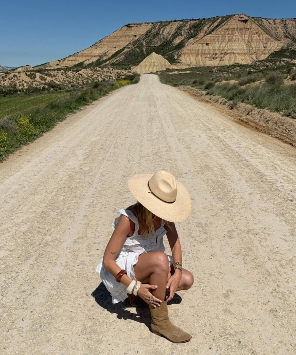 Pessoa agachada em estrada de terra, ajustando suas botas de cowboy. Montanhas no horizonte sob céu azul.