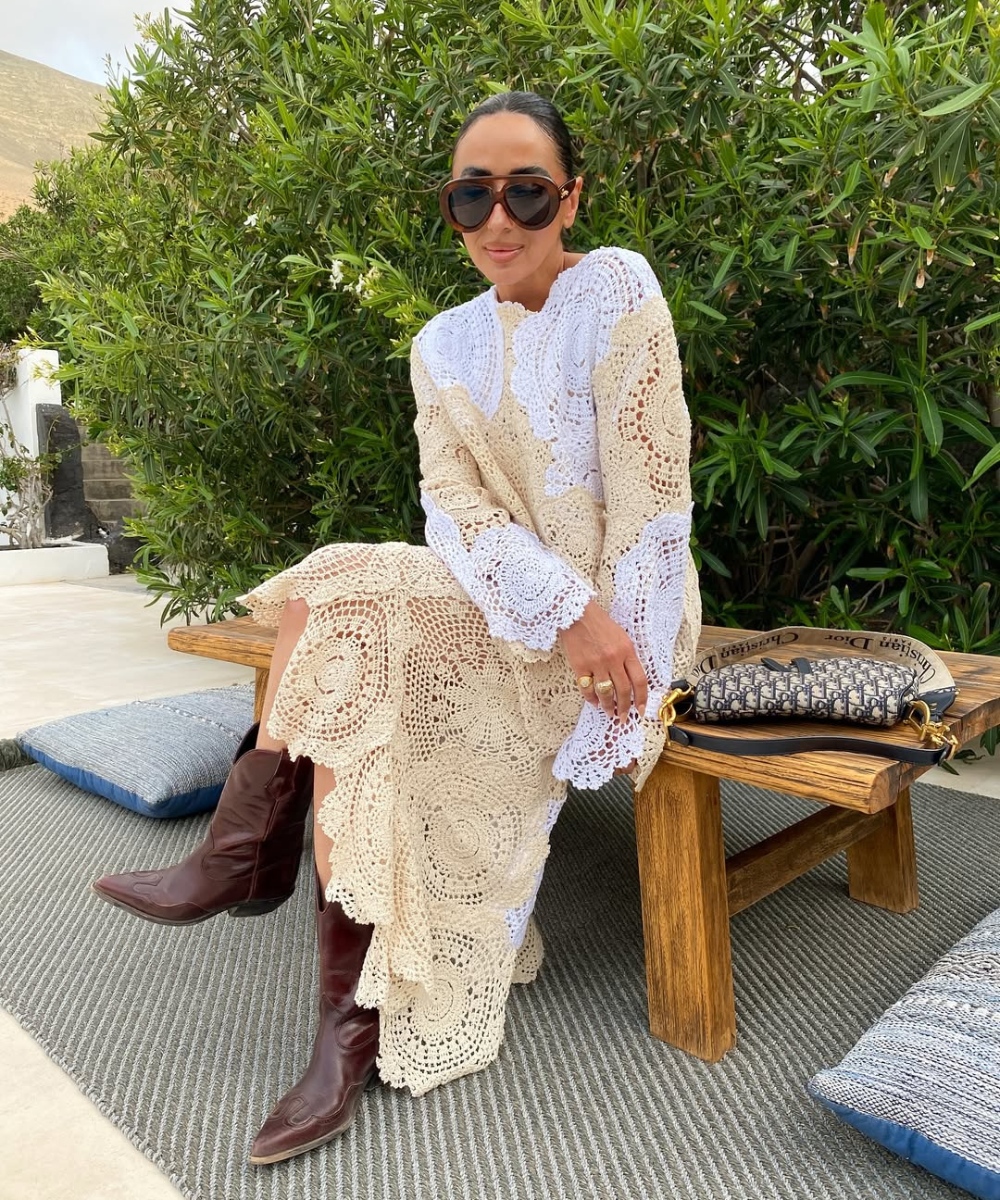 Mulher com vestido de crochê bege e botas de cowboy marrons sentada em um banco de madeira.