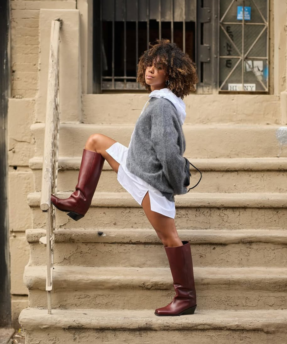 Pessoa com suéter cinza e vestido branco, usa botas marrons ao subir escadas.