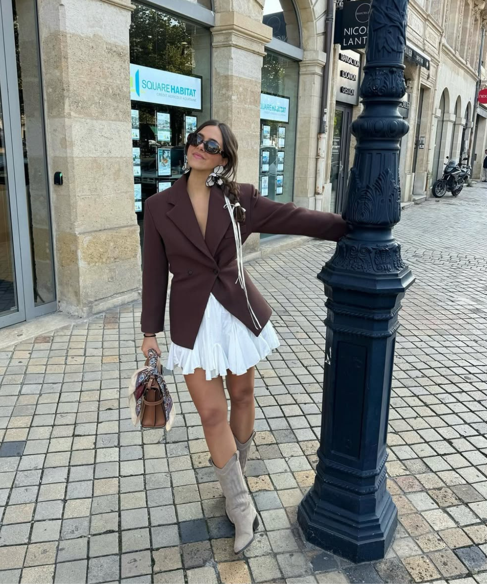 Pessoa em pé, vestindo blazer marrom e saia branca, junto a poste, usando botas de cowboy bege.