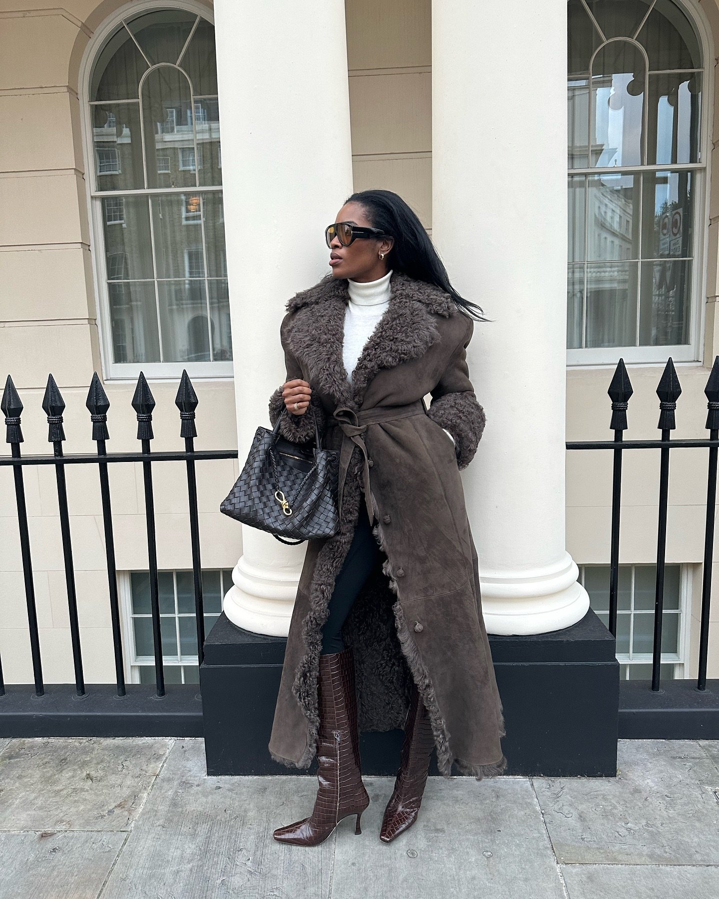 Mulher com casaco marrom e botas de bico fino marrons, segura uma bolsa grande.