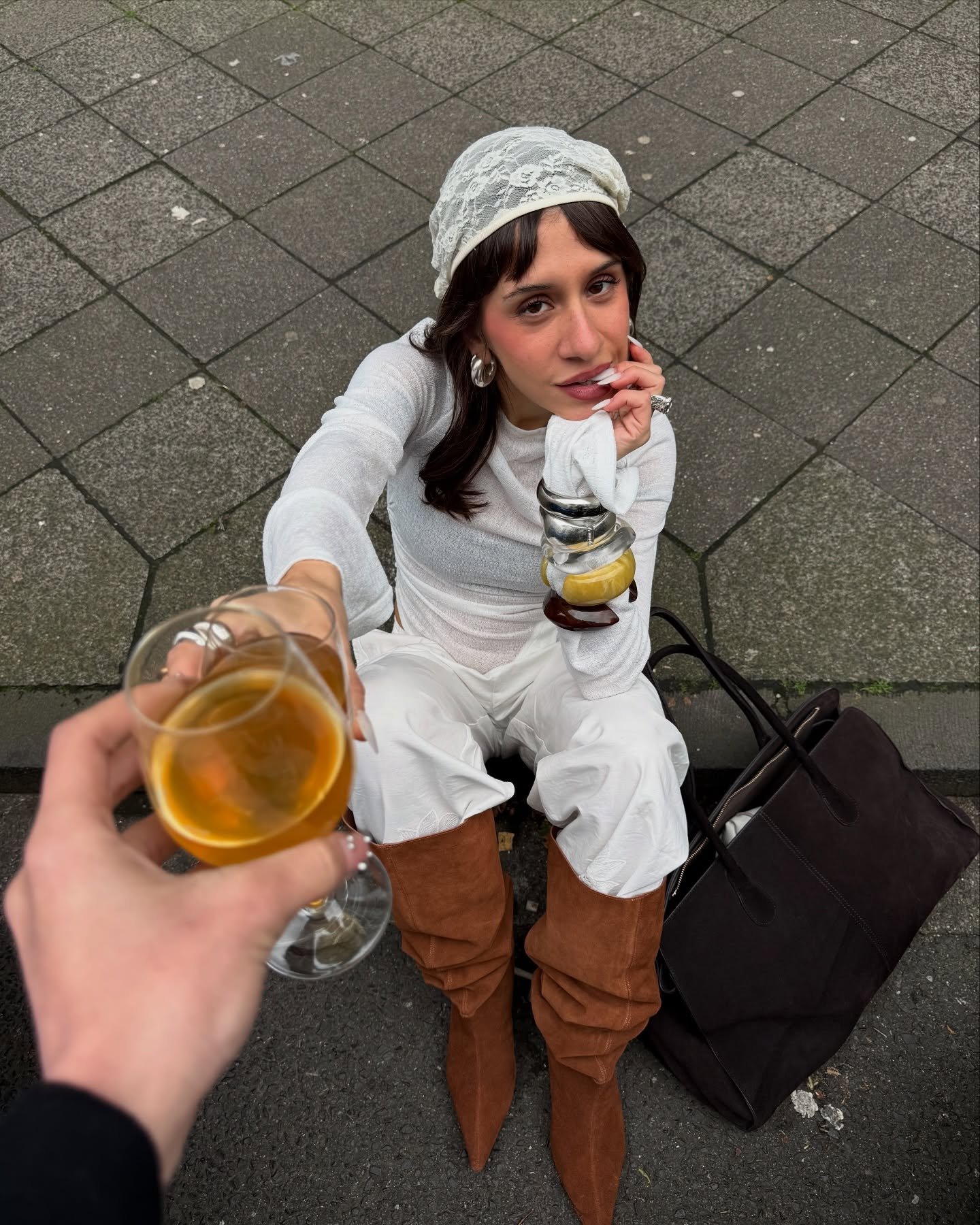 Pessoa sentada ao ar livre com botas de bico fino marrom, segurando um copo de bebida, acompanhada de uma bolsa preta.
