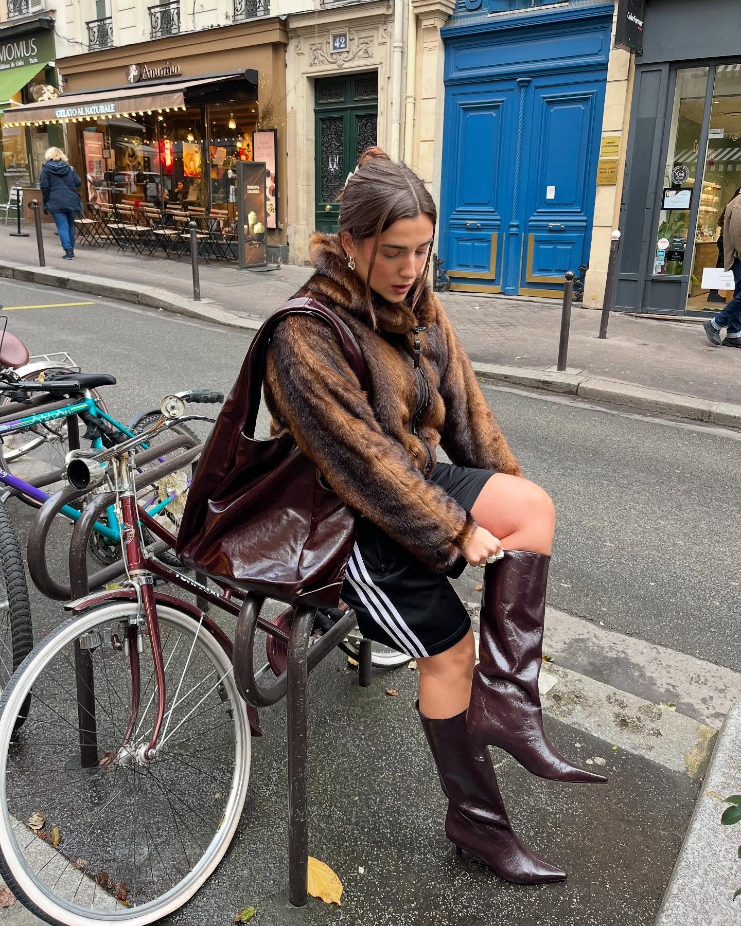 Mulher de casaco marrom sentada na calçada, ajustando botas de bico fino em ambiente urbano.
