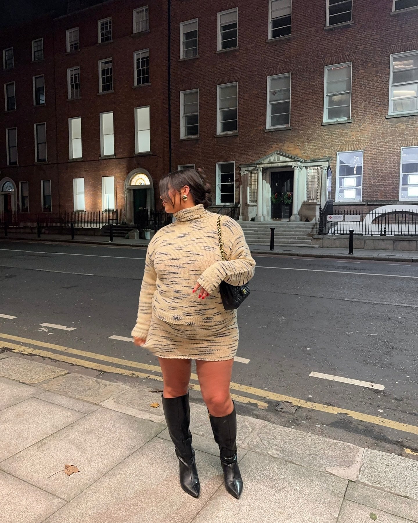 Mulher de vestido bege e botas de bico fino pretas em calçada urbana à noite.