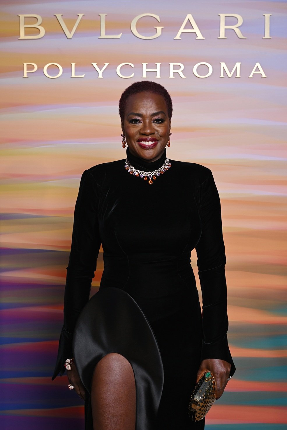 Pessoa sorridente em vestido elegante, fundo colorido de tons alaranjados e palavra Polychroma.