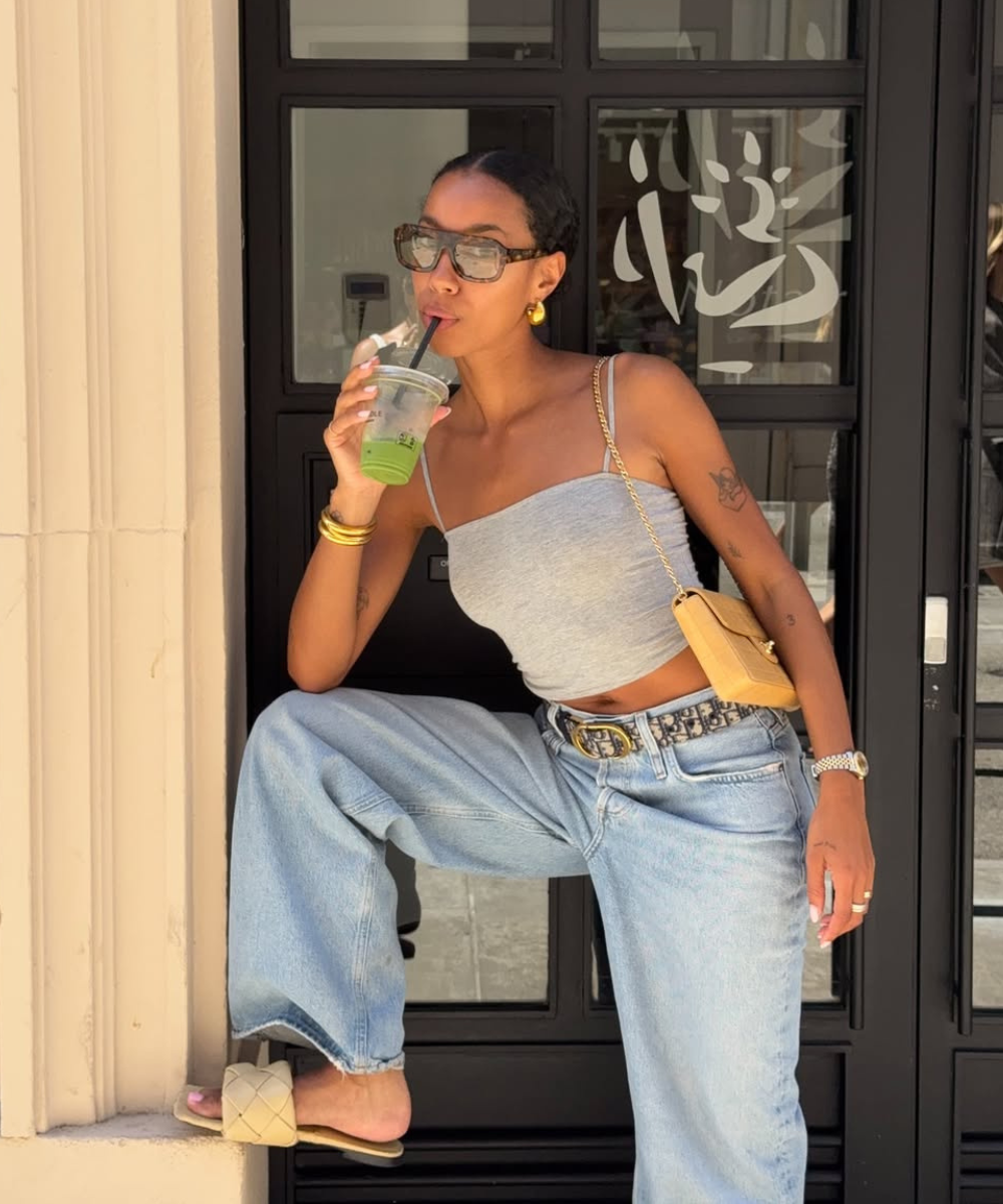 Pessoa com óculos de sol, calça jeans clara e top cinza, tomando bebida verde enquanto encosta em parede com porta preta.
