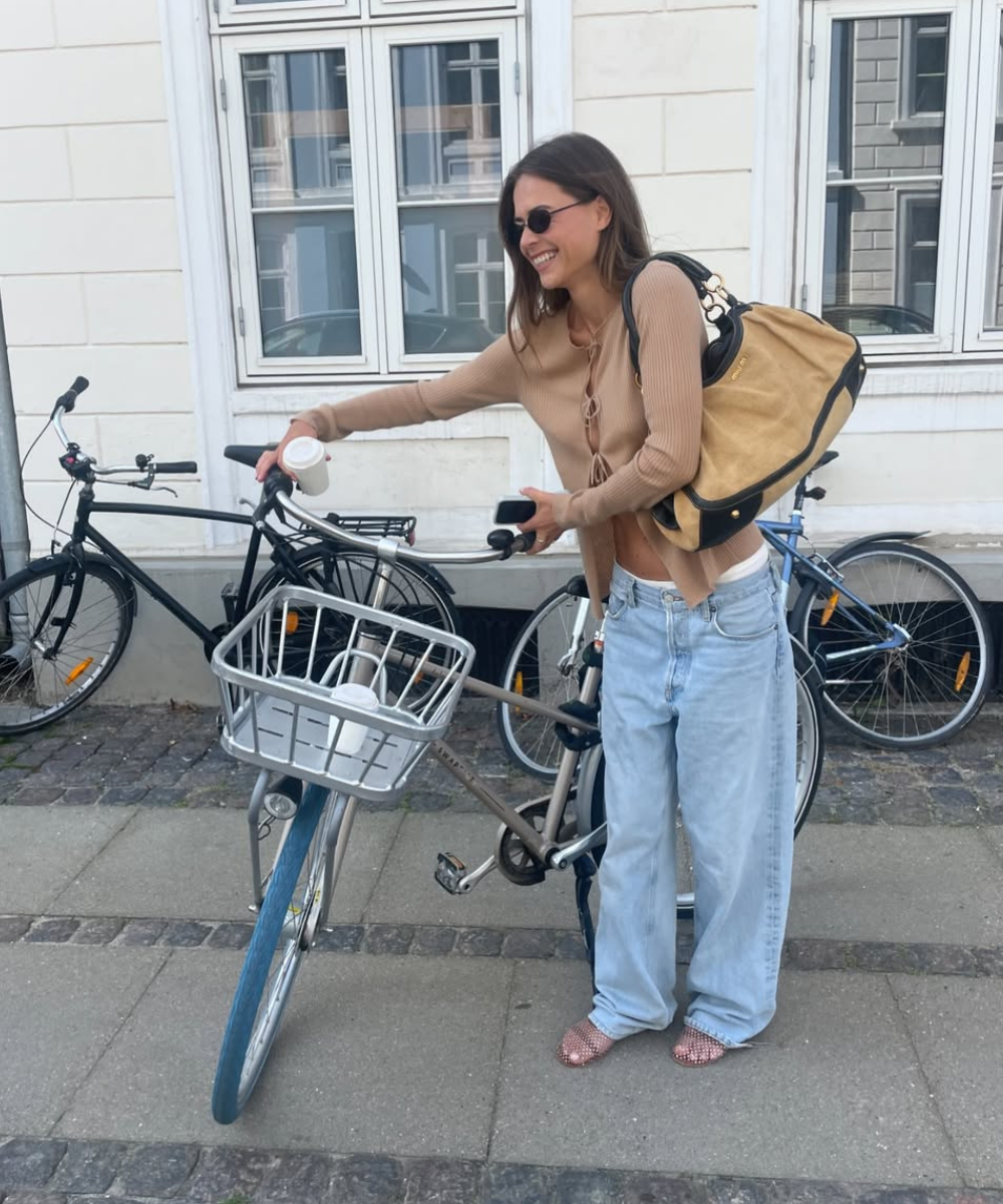 Pessoa sorrindo, usando calça jeans clara e blusa bege, segurando uma bicicleta na rua, com mochila no ombro.
