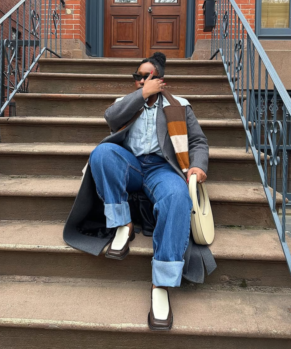 Pessoa sentada em escada vestindo calça jeans plus size, casaco cinza e lenço listrado, segurando bolsa clara.