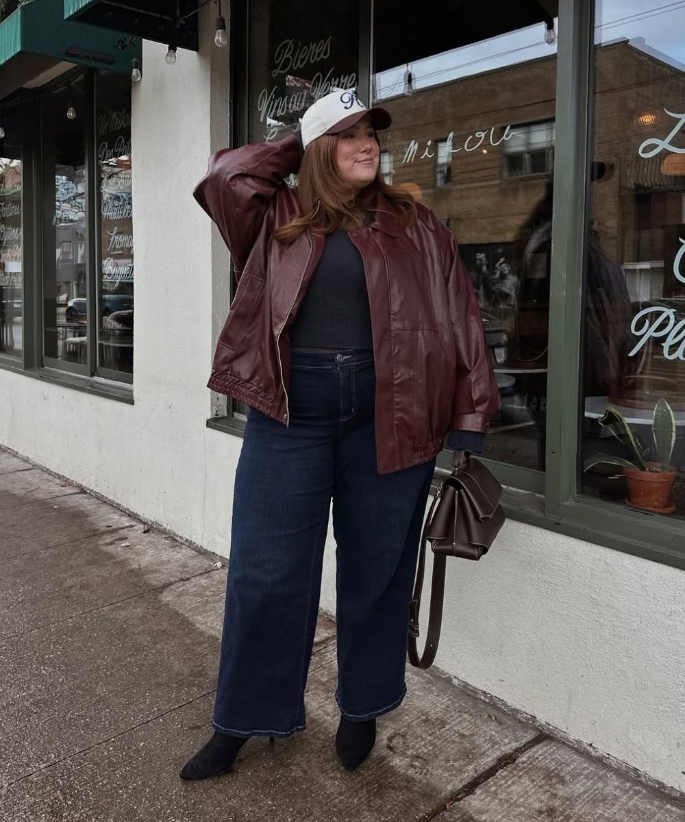 Usuária em frente a uma loja, usando jaqueta marrom, boné branco e calça jeans plus size. Carrega bolsa marrom.