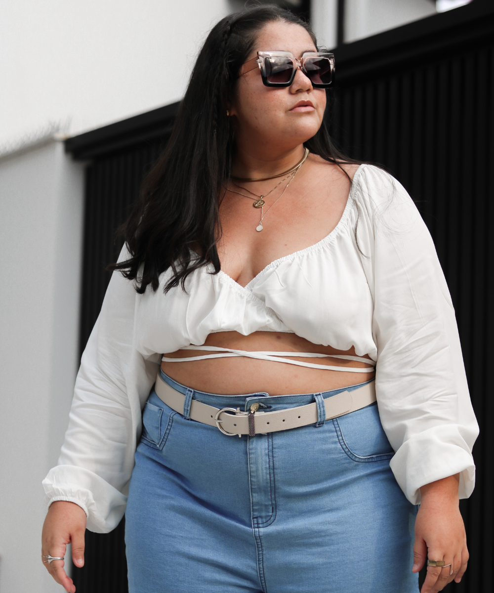 Mulher com óculos de sol vestindo blusa branca e calça jeans plus size, com cinto claro.