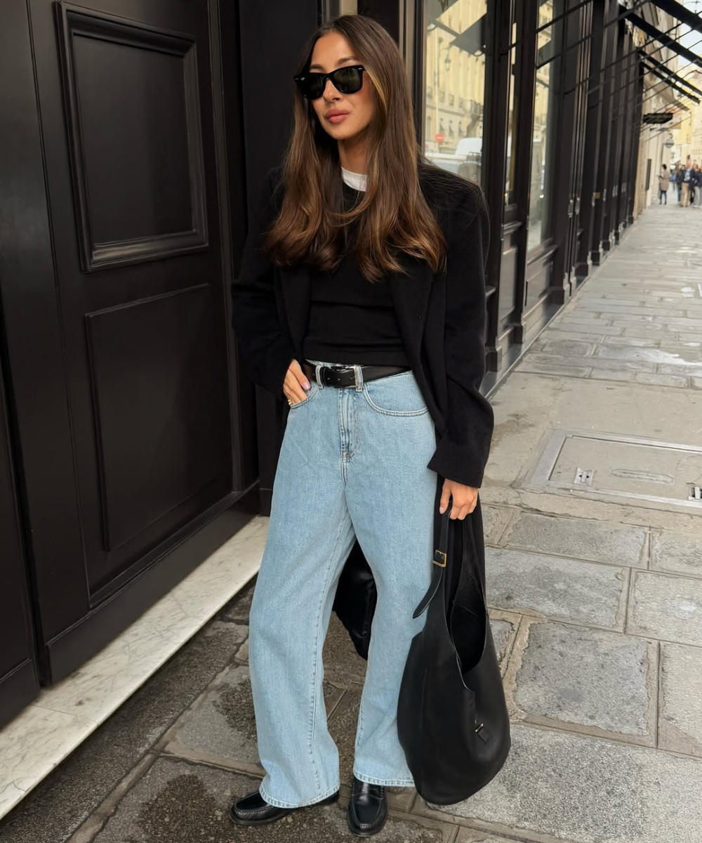 Mulher usando calças jeans largas, óculos escuros e casaco preto, com bolsa preta. Ela está em uma calçada.
