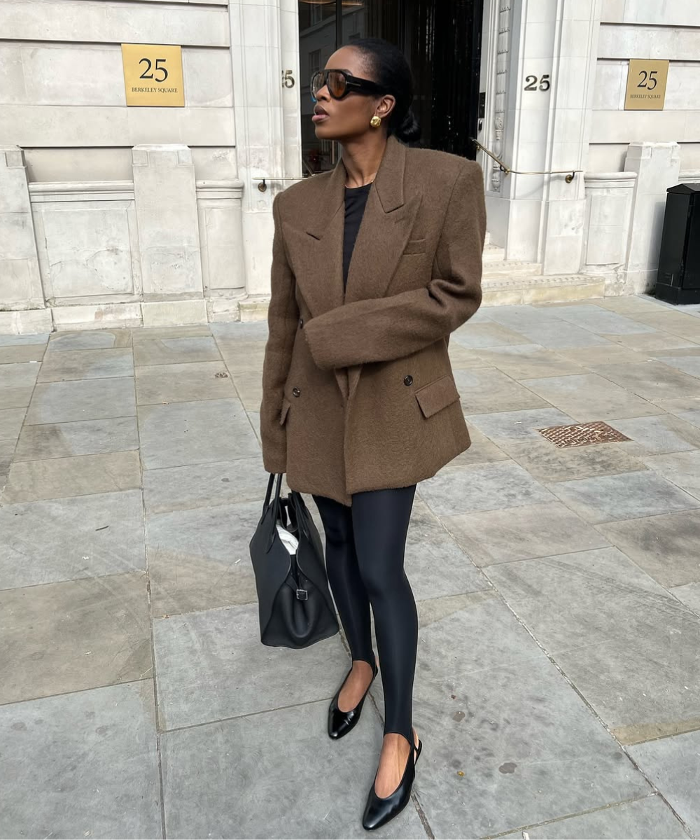 Pessoa veste blazer marrom e calça justa preta, usando bolsa preta e óculos escuros, em frente a um edifício com placas 25.