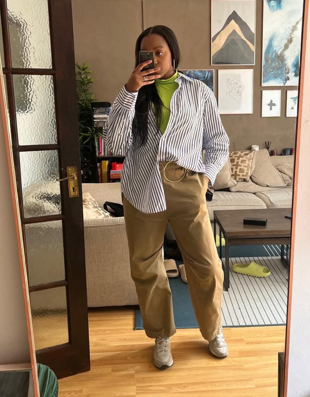 Pessoa tirando selfie usando camisa azul listrada e calça cáqui em sala decorada.