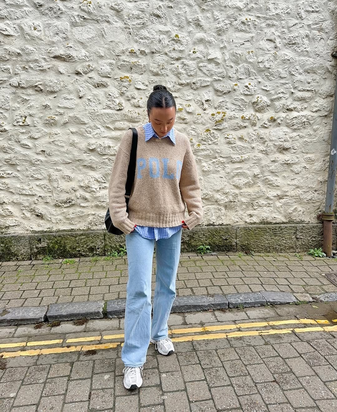 Pessoa de moletom bege e calça jeans, usando camisa azul listrada por baixo, em frente a uma parede de pedra.