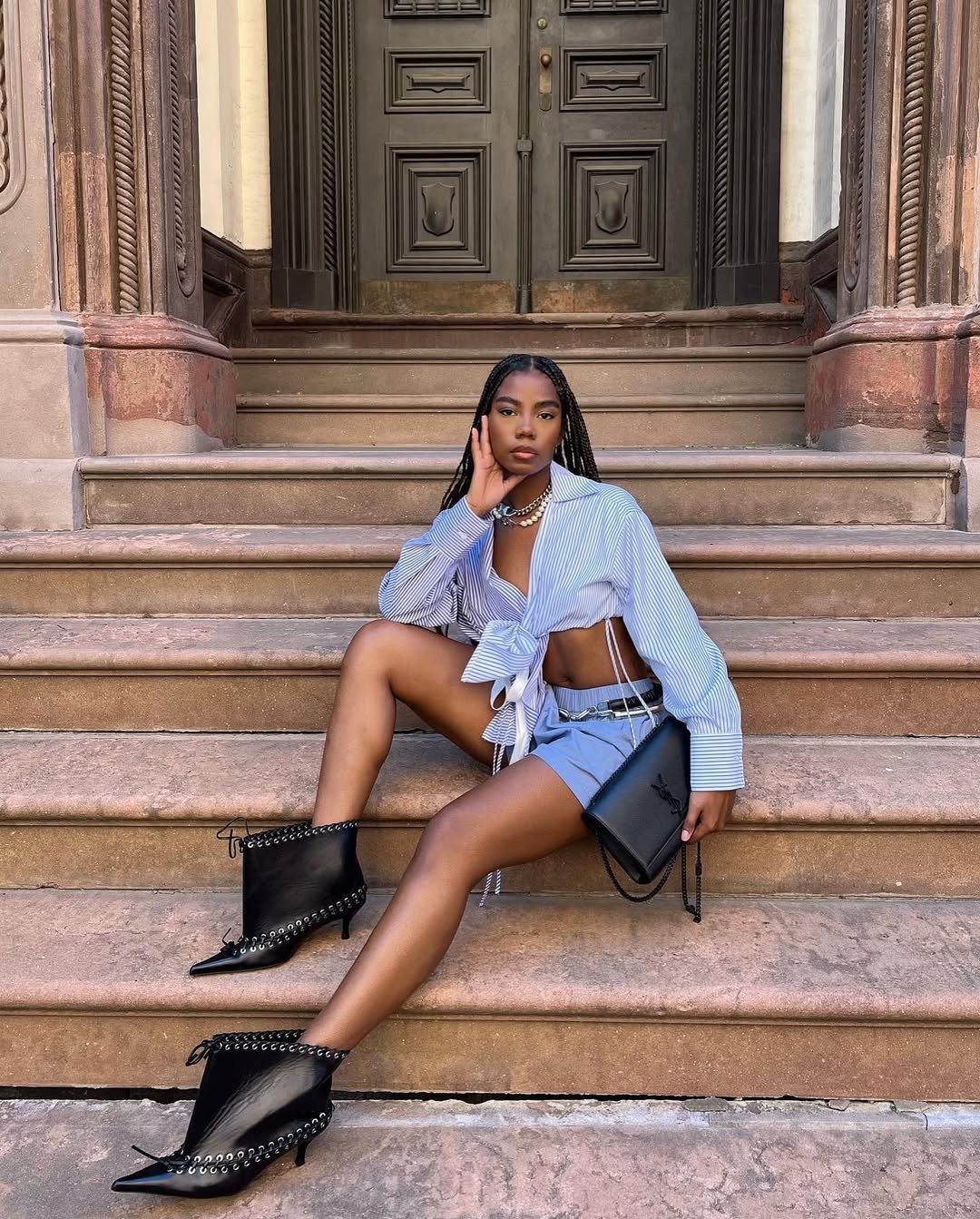 Pessoa com camisa azul listrada e shorts, sentada em escada de pedra. Usa botas pretas e segura uma bolsa preta.