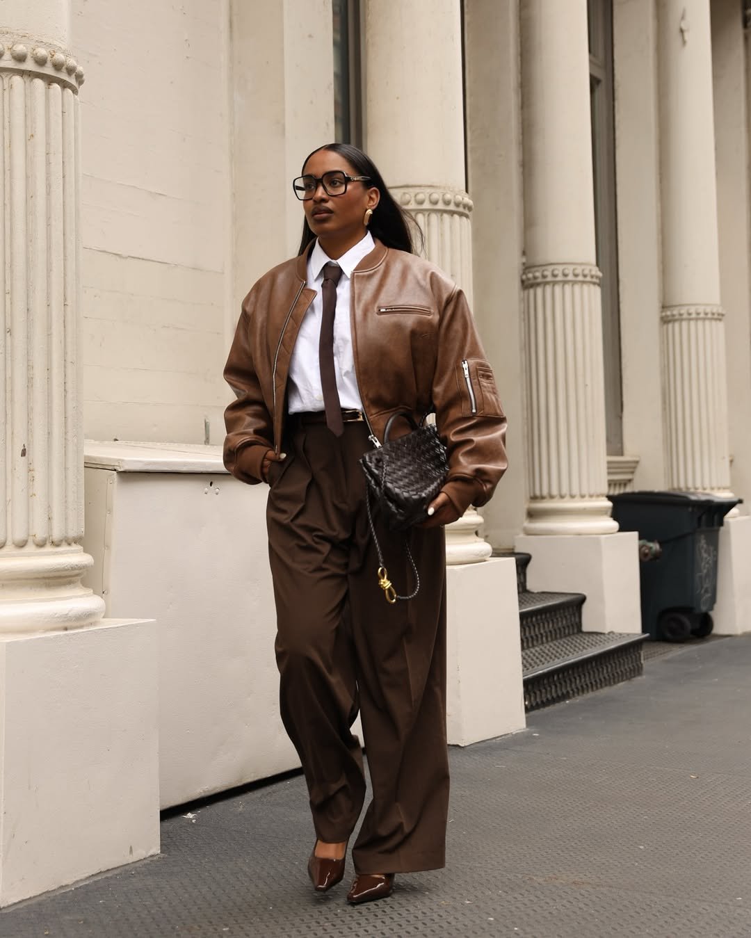 Mulher com calça e jaqueta marrom sob camisa branca feminina caminha por calçada urbana.
