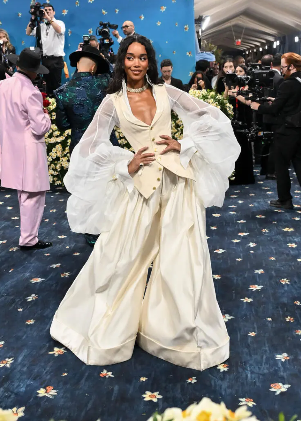 Vestido luxuoso com mangas bufantes e colar brilhante em evento elegante no tapete azul, rodeado por fotógrafos.