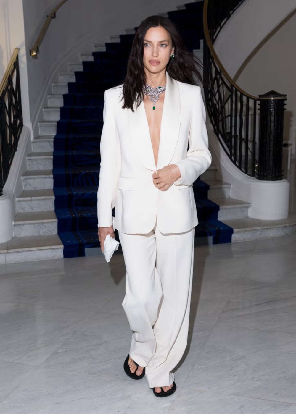 Modelo usando terninho branco elegante e colar luxuoso, descendo escada de mármore com guarda-corpo curvo.