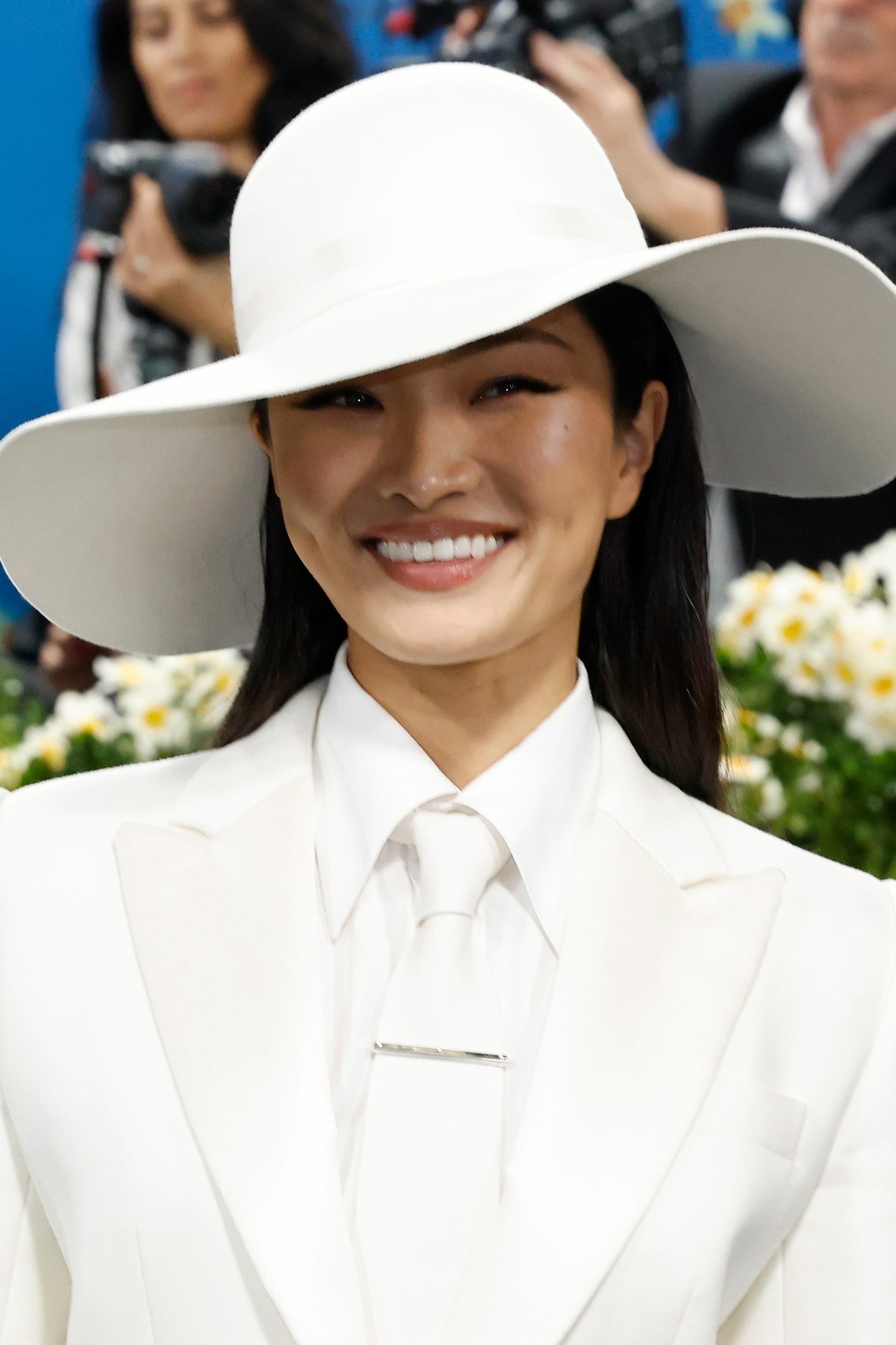 Pessoa sorridente usando chapéu e roupa branca elegante. Estilo associável à tendência do Met Gala 2025.