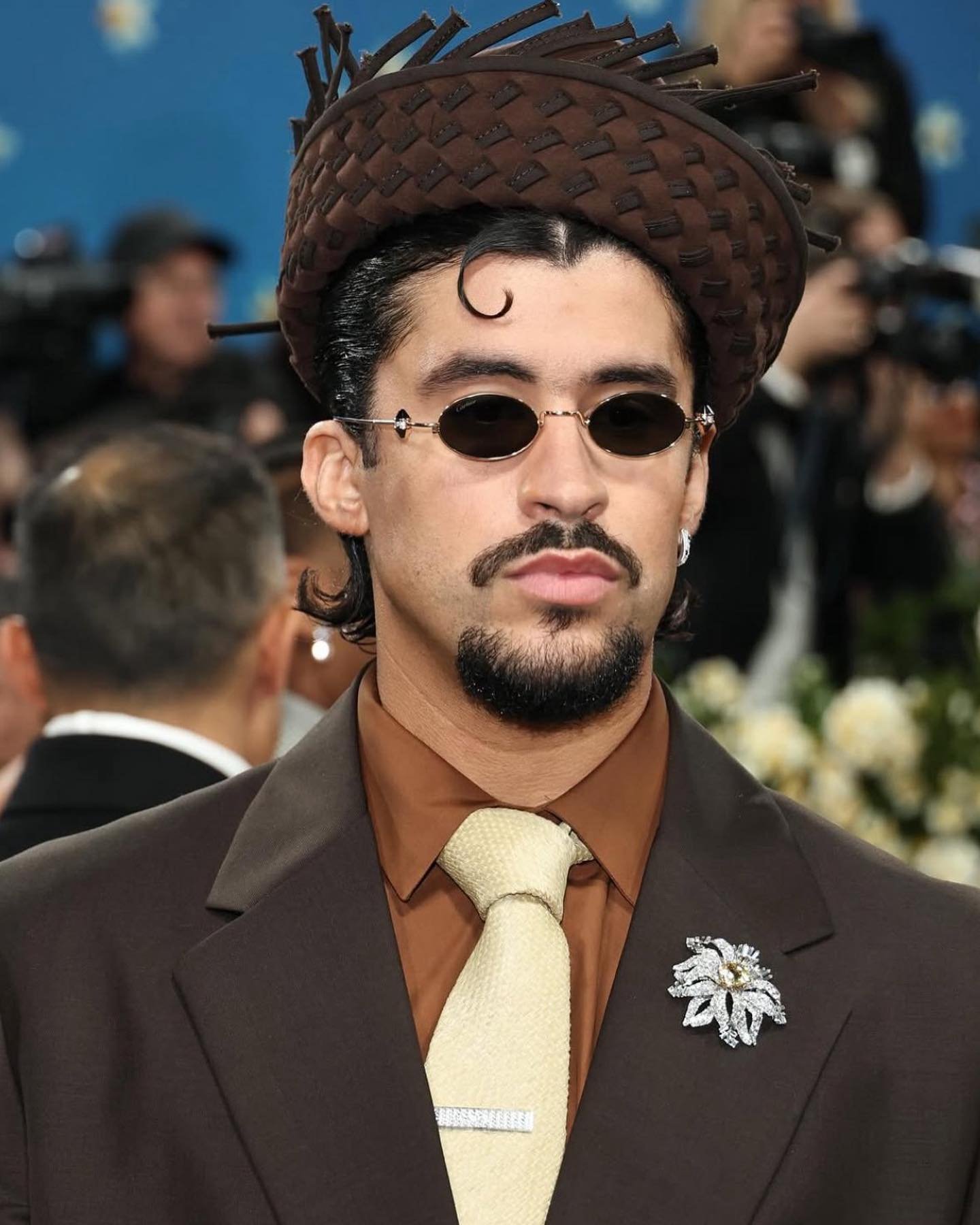 Homem usando terno marrom com broche prateado no Met Gala; chapéu de design inovador, explorando tendência de 2025.