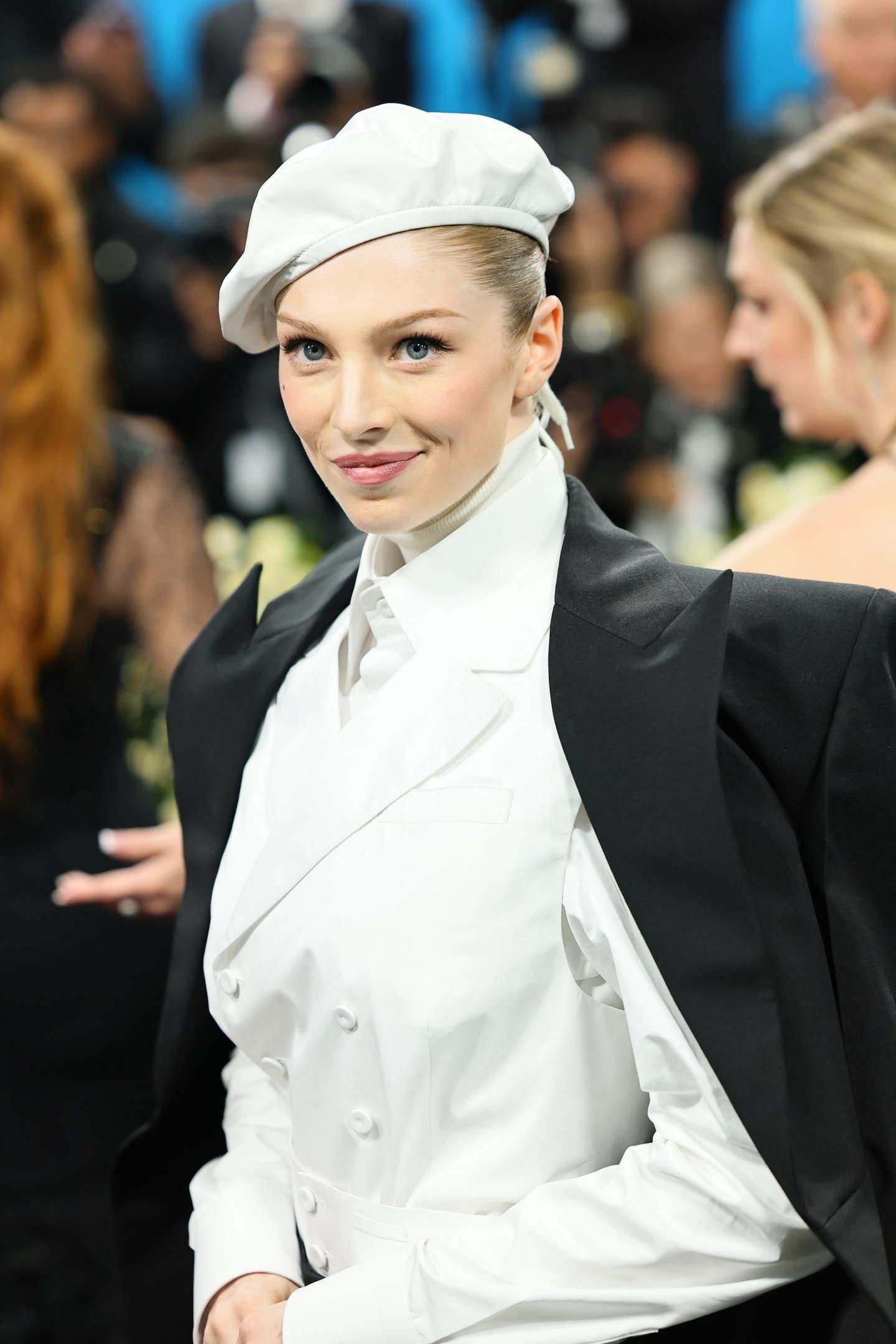 Pessoa vestindo traje elegante com boina branca e paletó preto, em ambiente de evento glamuroso.