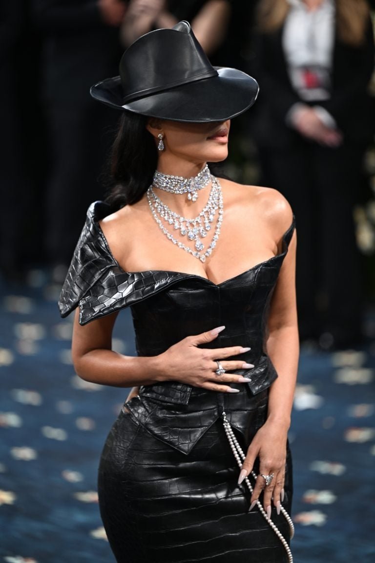 Mulher em vestido preto de couro com decote, colares brilhantes e chapéu. Elegância futurista, possivelmente tendências de moda 2025.