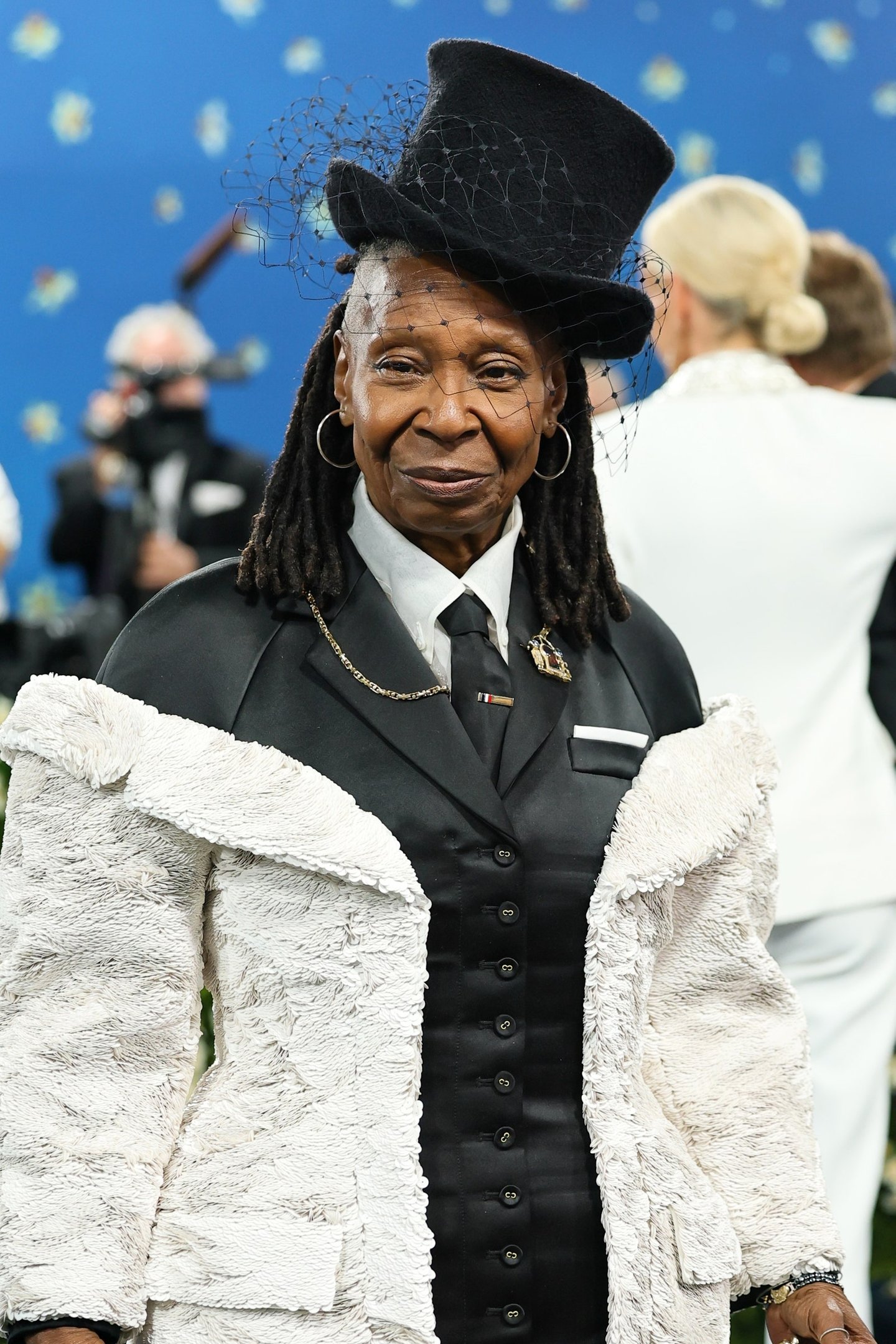 Pessoa em traje elegante, terno preto e casaco texturizado, chapéu preto decorado, possível moda do Met Gala 2025.