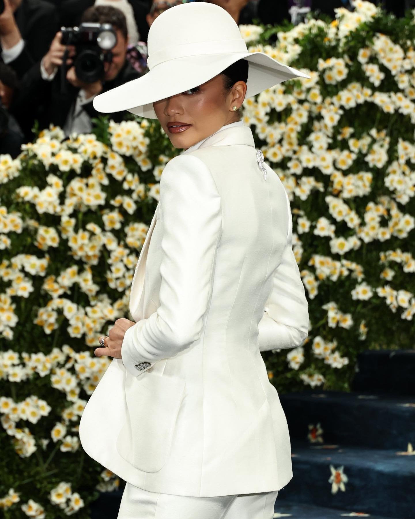 Pessoa em terno branco e chapéu largo, posando em evento elegante com fundo florido.