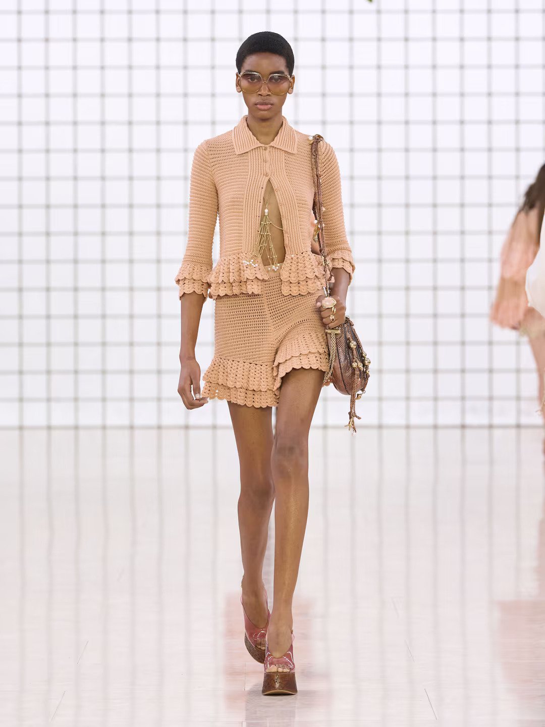 Modelo desfila com conjunto de crochê bege e acessórios, destacando tendências de beleza atuais.