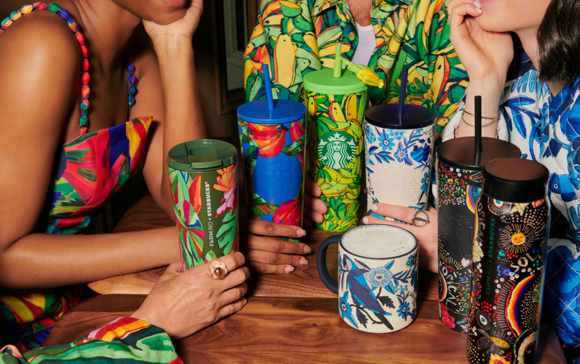 Pessoas segurando copos e caneca Starbucks com estampas coloridas, sentadas à mesa.