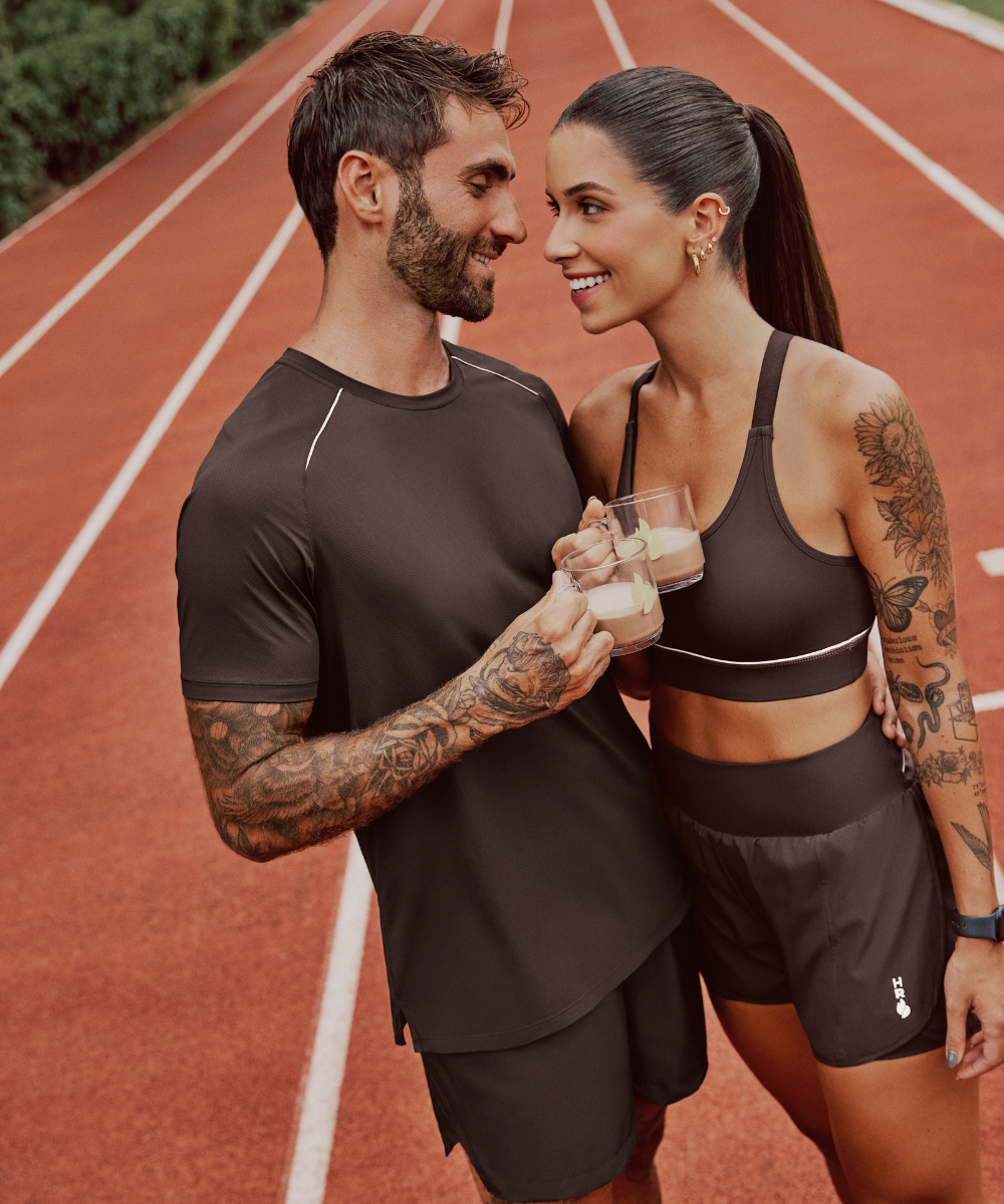 Casal sorridente em pista de corrida, segurando copos com bebida, vestindo roupas esportivas. Collab Caffeine Army.