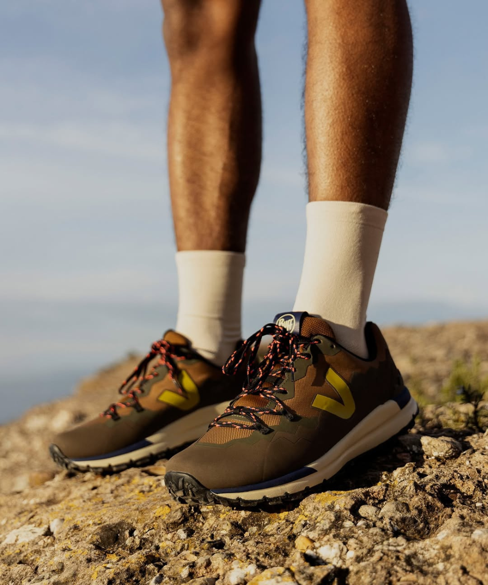 Tênis para trilhas em terreno rochoso, ideal para hiking, com meias brancas e céu ao fundo.