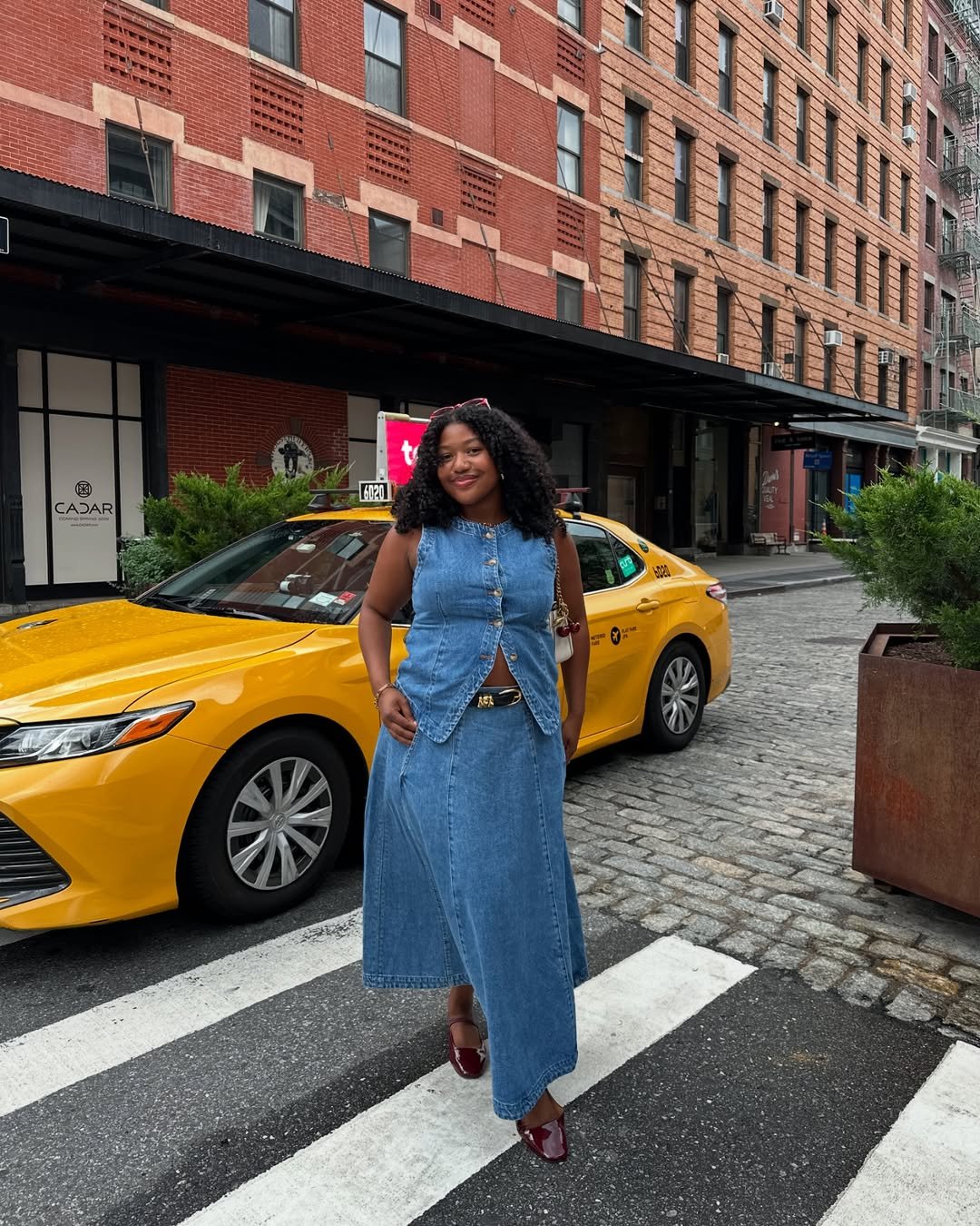 Usuária com vestido jeans e sapatilhas marrons diante de táxi amarelo em rua urbana.