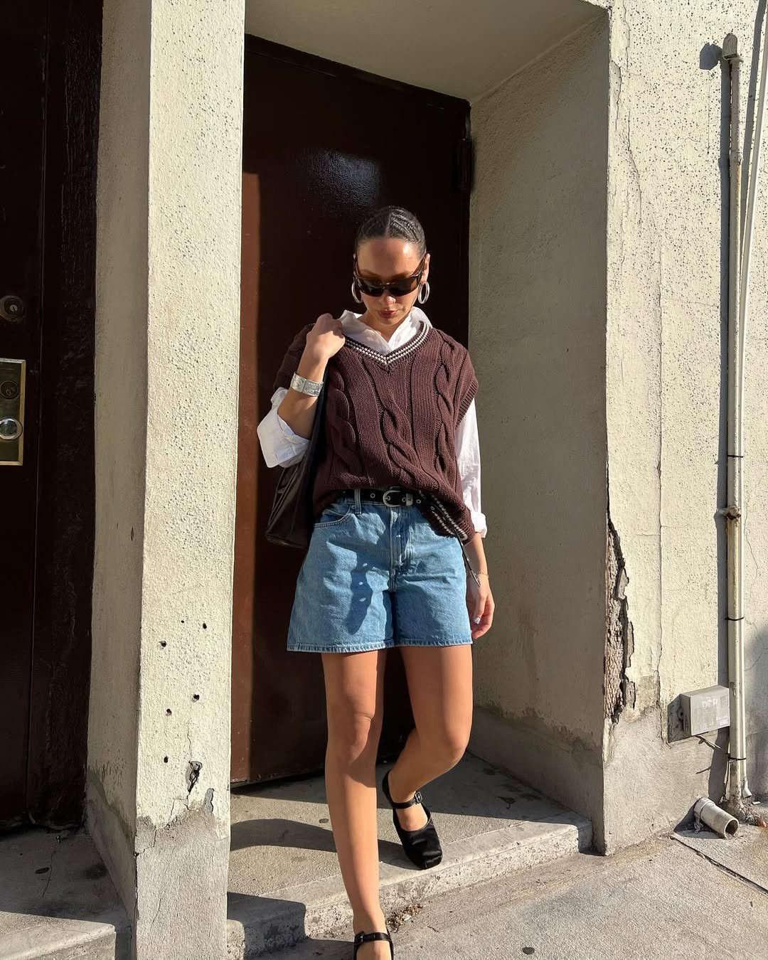 Pessoa com shorts jeans, colete marrom e sapatilha preta, vestindo camisa branca e óculos escuros.