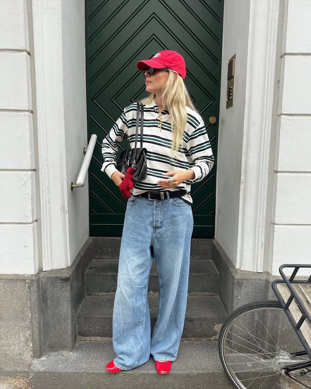 Pessoa com boné vermelho veste jeans largos e sapatilhas vermelhas, próximo a uma porta verde.