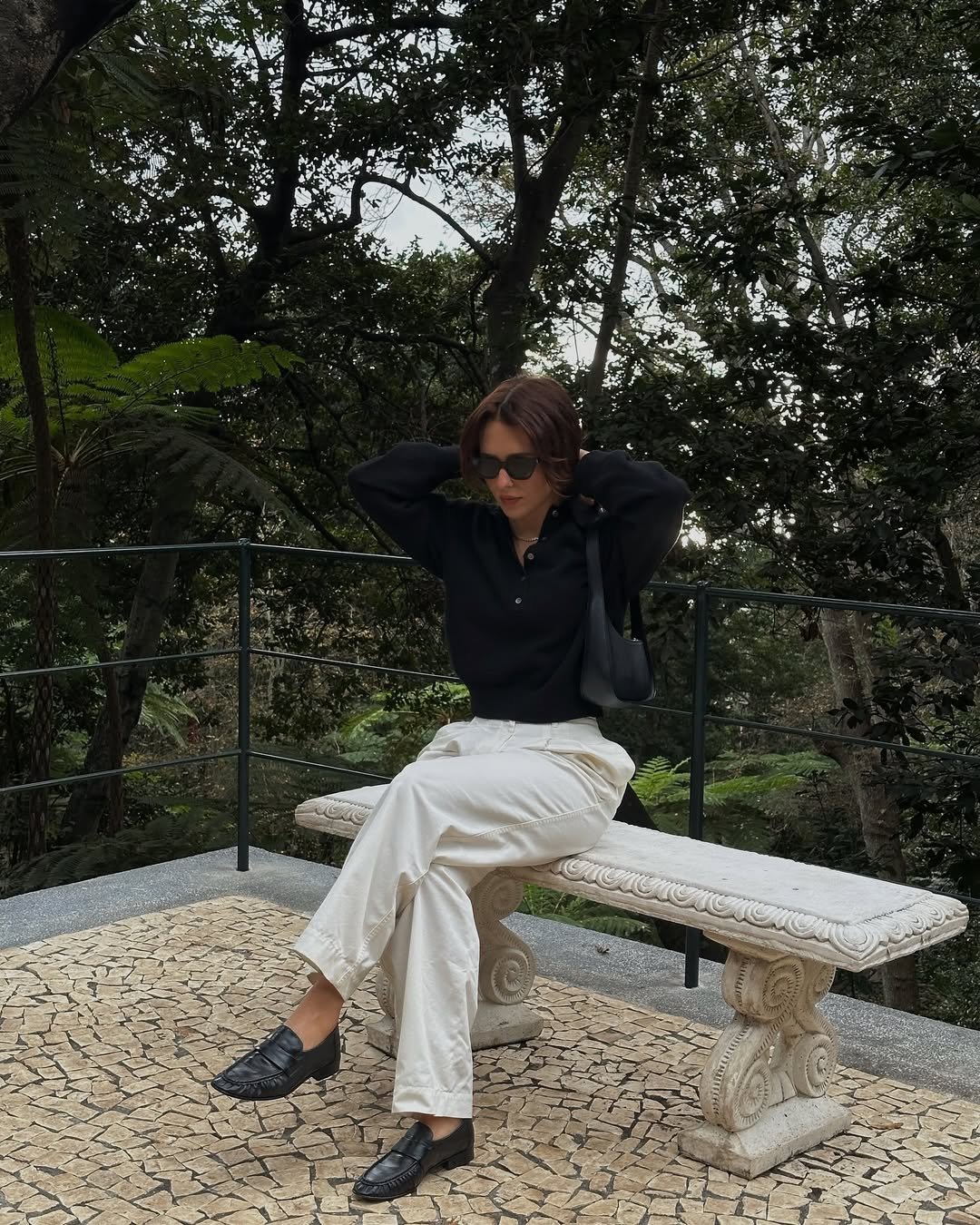 Pessoa sentada em banco de pedra, usando camisa preta, calça branca e loafers pretos, em área arborizada.