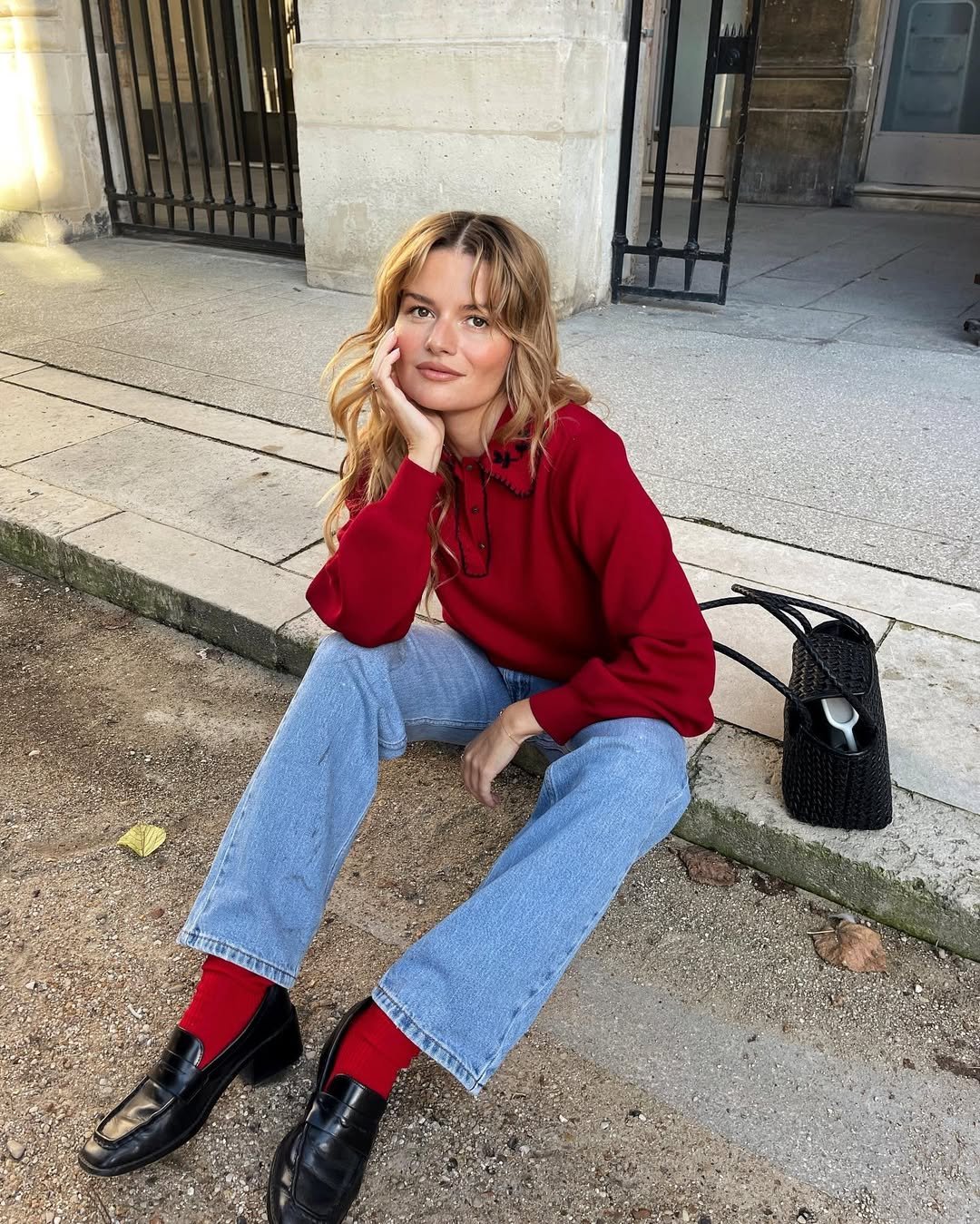 Pessoa sentada no chão, usando suéter vermelho, calça jeans e loafers pretos. Bolsa preta ao lado.