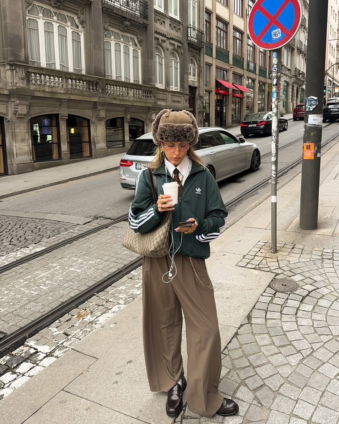 Pessoa com casaco verde e chapéu de pele falsa, segura café na rua; usa calças largas e loafers marrons.